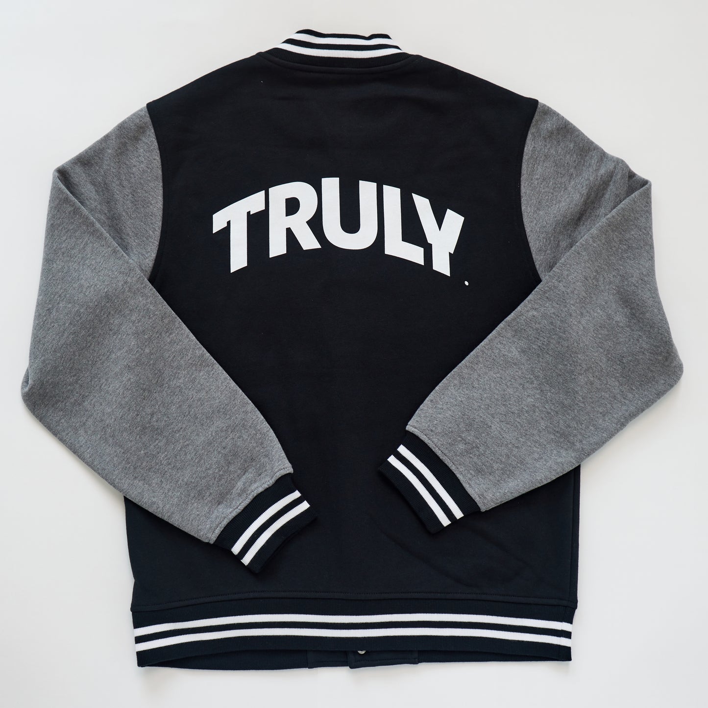 A bomber jacket with a dark gray body and light gray sleeves, viewed from the back. The word "TRULY" is emblazoned in large, white, bold letters across the upper back. The collar, cuffs, and waistband are black with two parallel white stripes.
