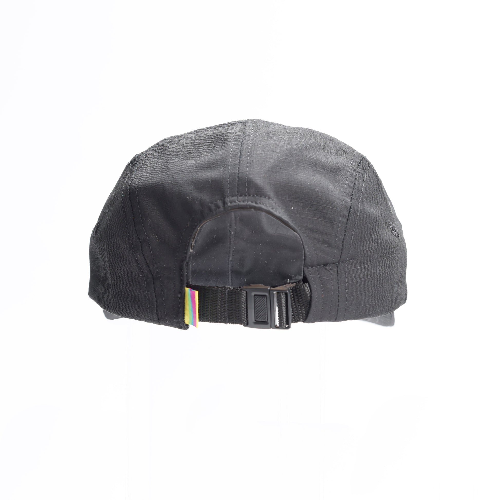 The back view of a black five-panel baseball cap, showing an adjustable strap with a black buckle and a colorful woven tag on the side.