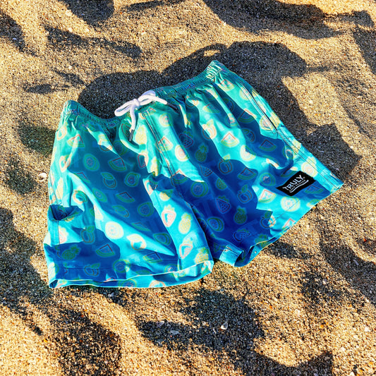 A pair of bright blue swim trunks with a pattern of light blue and green abstract fruit shapes, lying on sun-drenched sand. A small black and white tag with the "TRULY" logo is visible on the right leg.