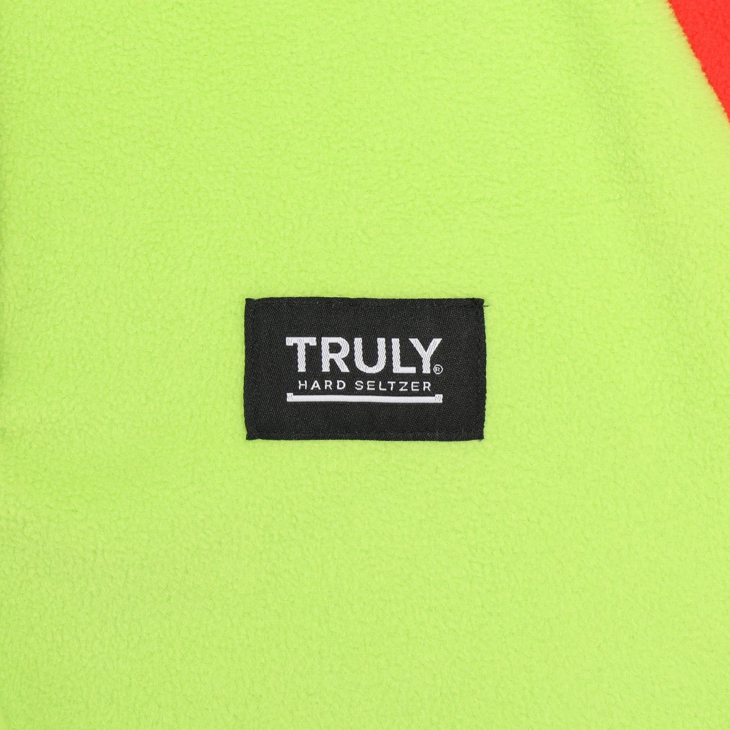 A close-up of a neon yellow fleece fabric with a black rectangular patch in the center. The patch has "TRULY." in white bold letters with "HARD SELTZER" and a horizontal line underneath.