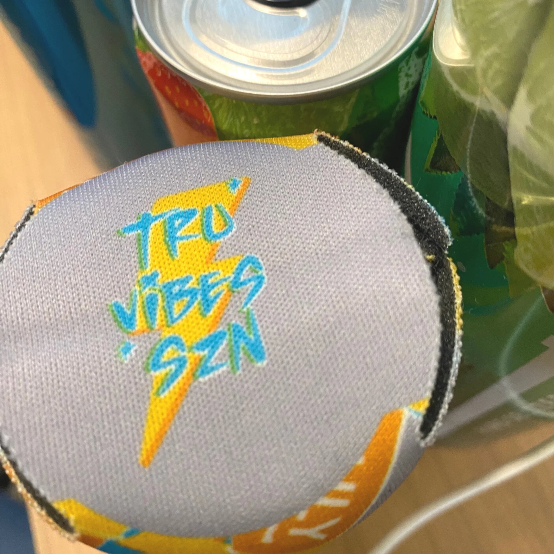 A close-up, top-down view of the bottom of a slim can cooler, revealing "TRU VIBES SZN" with a lightning bolt graphic in blue and yellow on a light gray background. Parts of two other Truly seltzer cans are visible in the blurry background.