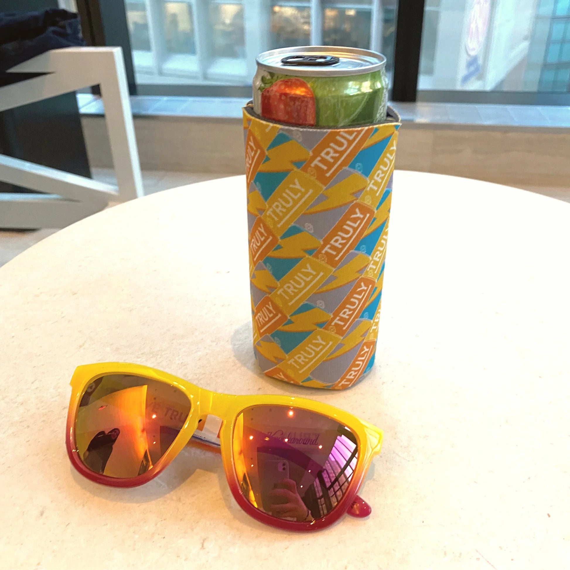 A light white table with a light yellow to red ombre pair of Knockaround sunglasses in the foreground. In the background, a light gray slim can cooler with yellow, blue, and gray repeating "TRULY" logos and lightning bolts holds a Truly seltzer can.