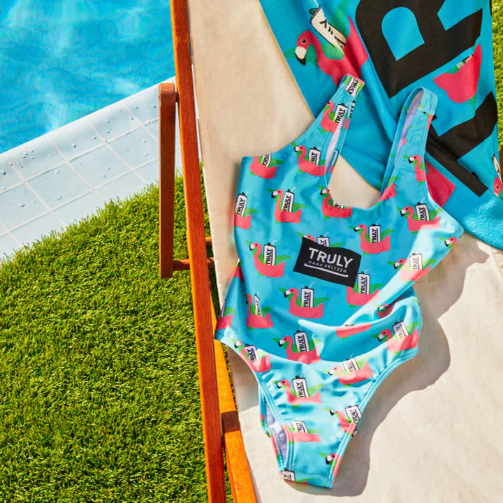 A light blue one-piece swimsuit with a repeating pattern of pink flamingos, each holding a white Truly Hard Seltzer can. A large black patch with "TRULY HARD SELTZER" in white text is centered on the chest. The swimsuit is draped over a light-colored lounge chair next to a swimming pool and green grass.