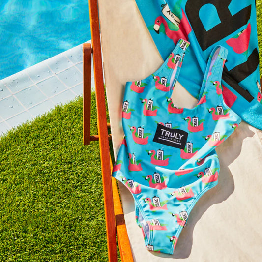 A light blue one-piece swimsuit with a repeating pattern of pink flamingos, each holding a white Truly Hard Seltzer can. A large black patch with "TRULY HARD SELTZER" in white text is centered on the chest. The swimsuit is draped over a light-colored lounge chair next to a swimming pool and green grass.