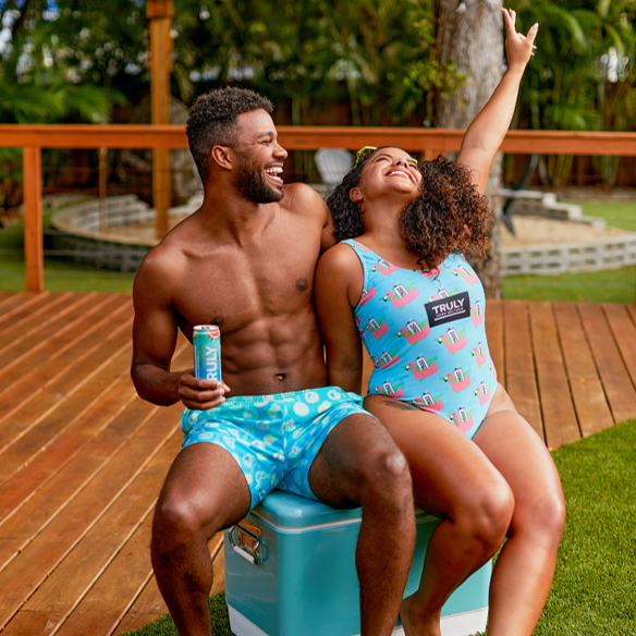 A man with a beard and a shirtless, torso sits on a teal and white cooler, holding a Truly Hard Seltzer can. He is looking at and laughing with a woman to his right who has curly hair and is wearing a light blue swimsuit with a flamingo and "TRULY" pattern. She is raising her right arm in the air and looking up. They are outdoors on a patio with wooden decking and grass in the background.