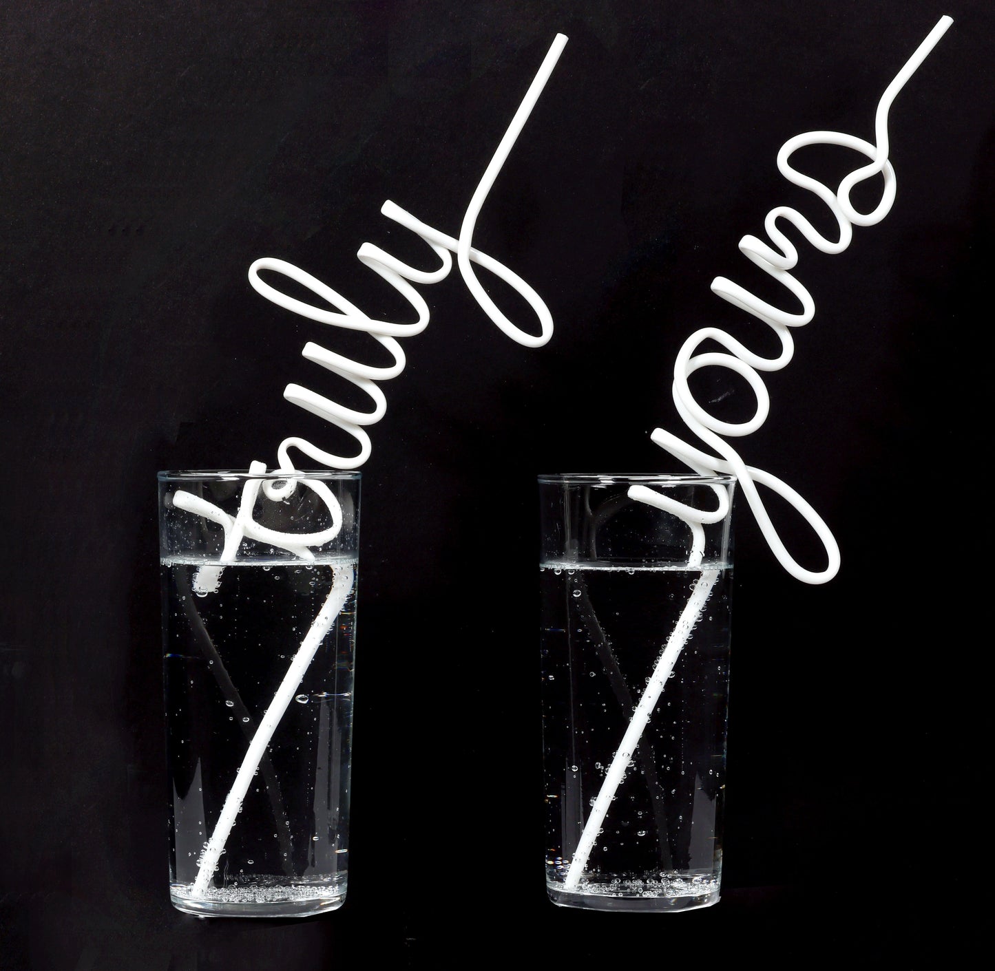 Two clear glasses filled with clear liquid, each containing a white novelty straw that spells out a word. The straw in the left glass spells 'truly', and the straw in the right glass spells 'yours'. Both straws extend diagonally upwards from the glasses. The background is black.