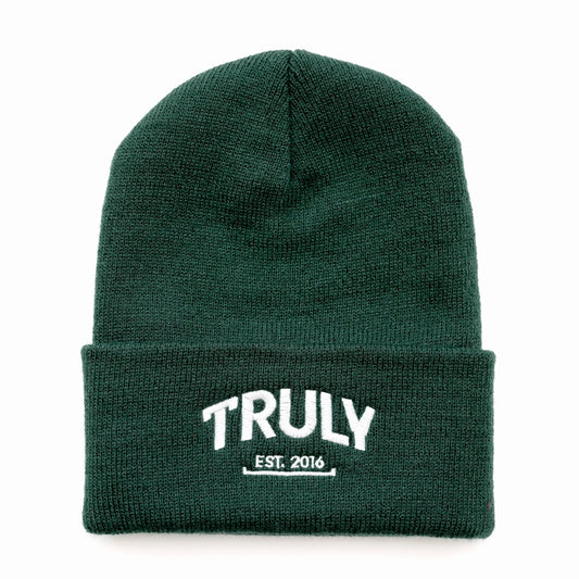 A dark green knit beanie with a folded cuff. "TRULY" is embroidered in white letters with "EST. 2016" underneath, also in white.