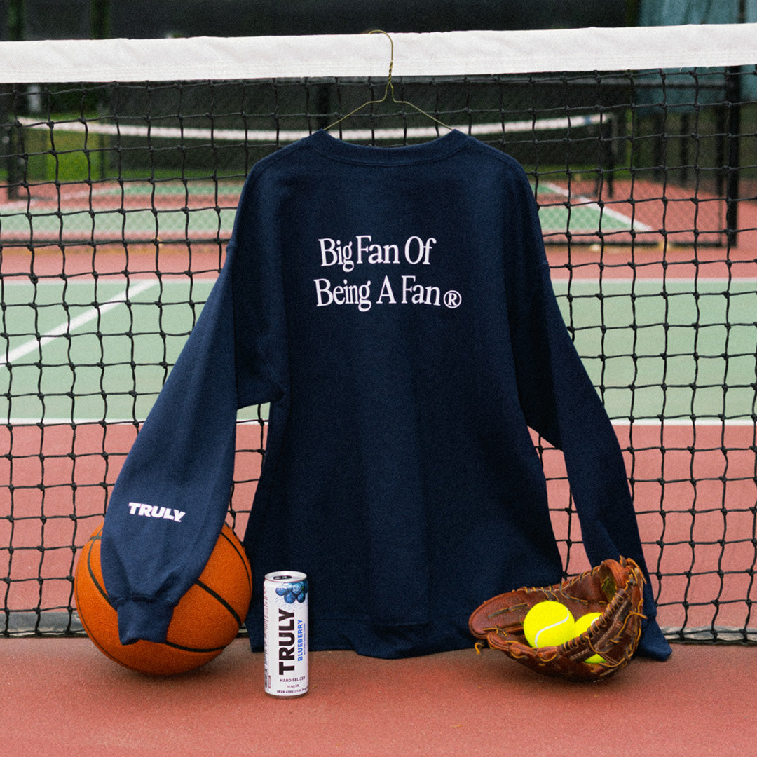 A navy blue long-sleeve sweatshirt with white text reading 'Big Fan Of Being A Fan' is displayed on a hanger on a tennis court net, styled with a basketball, a can of TRULY soda, and a baseball glove with tennis balls.
