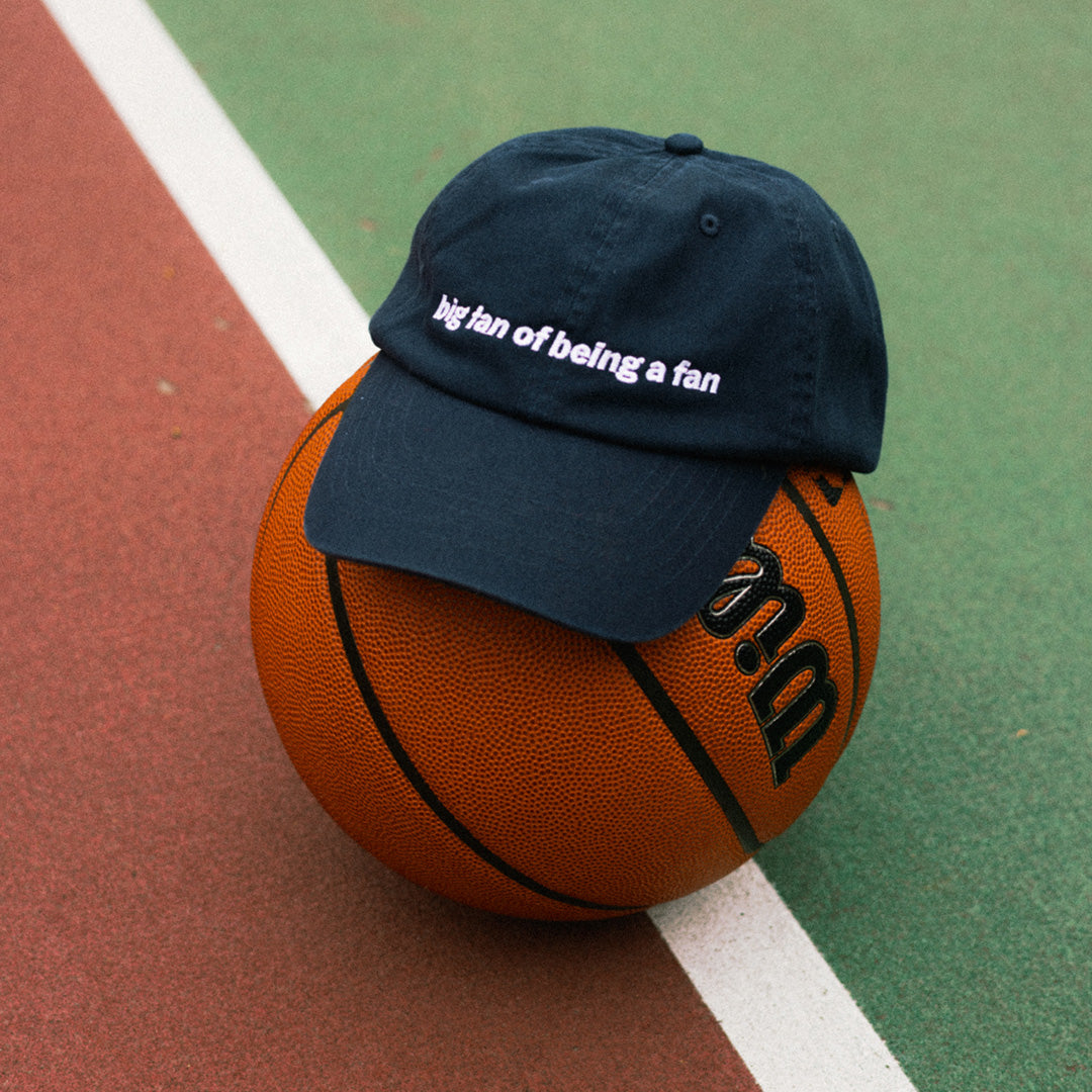 A navy blue baseball cap with white embroidered text reading 'big fan of being a fan' is placed on top of an orange basketball on an outdoor court with green and red surfaces and a white line.
