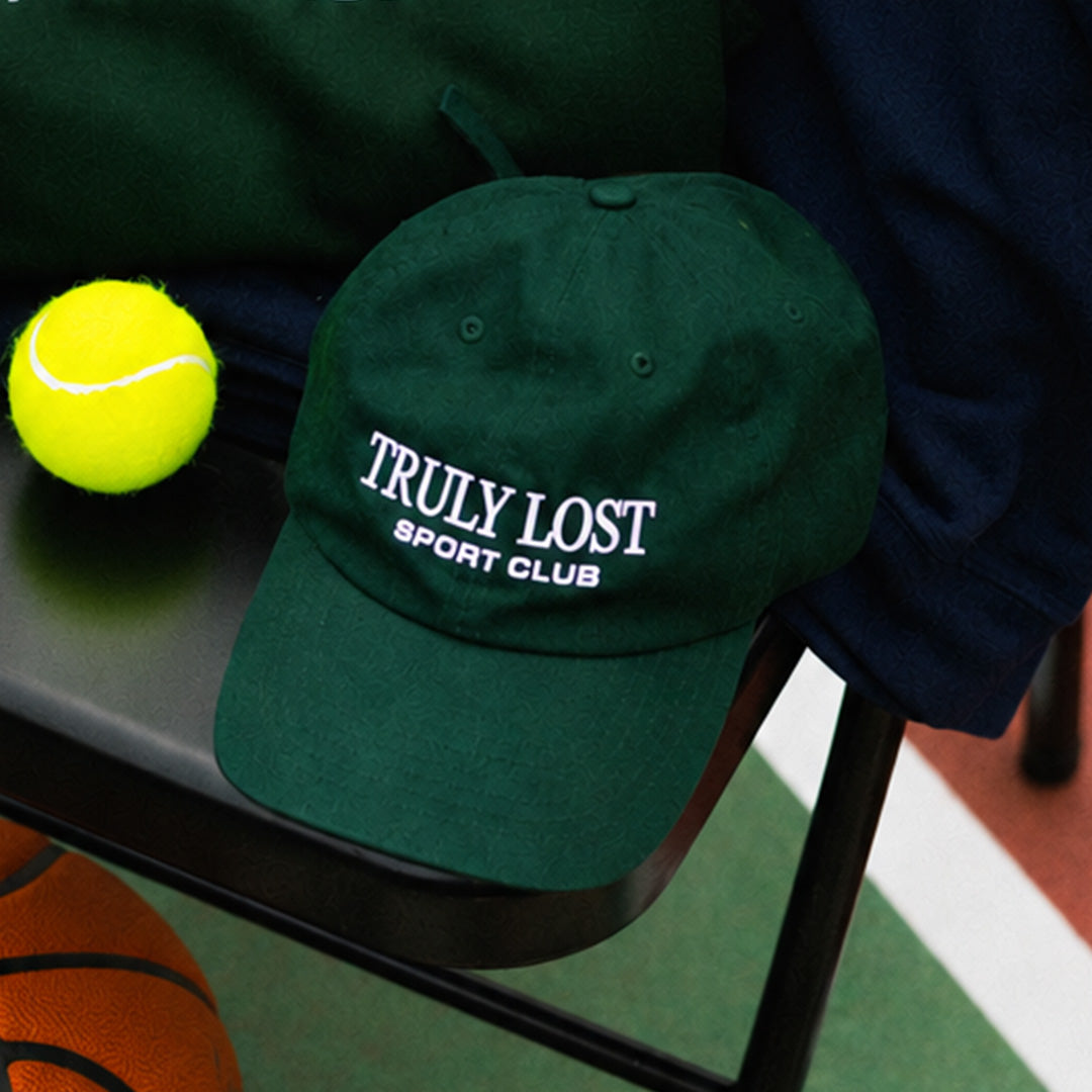 A dark green baseball cap with 'TRULY LOST SPORT CLUB' embroidered in white on the front, resting on a black bench next to a yellow tennis ball.