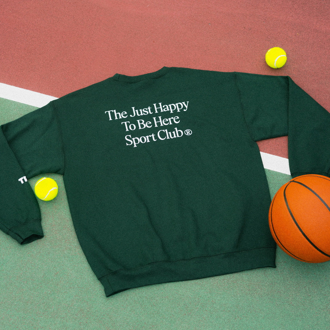 A dark green crewneck sweatshirt with white text reading 'The Just Happy To Be Here Sport Club™' on the back, styled flat-lay on a tennis court with tennis balls and a basketball.