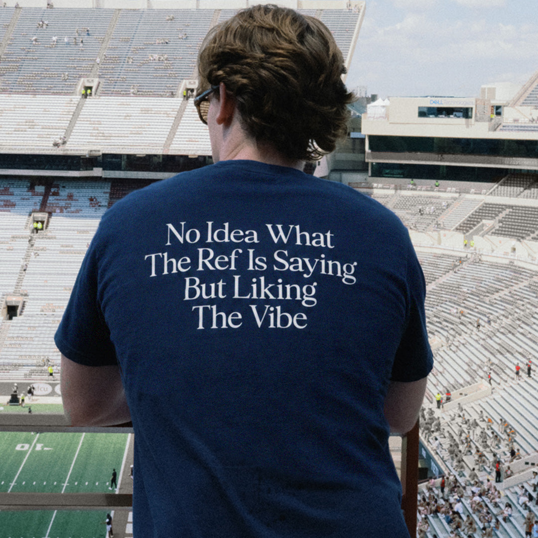 A person seen from behind wearing a navy blue t-shirt with white text that reads 'No Idea What The Ref Is Saying But Liking The Vibe', standing in a large outdoor stadium overlooking the field and bleachers.
