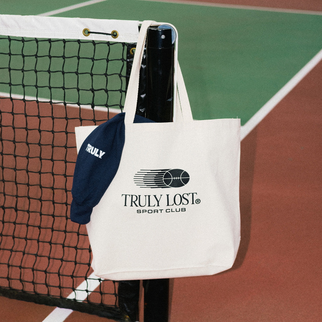 A cream canvas tote bag with black 'TRULY LOST SPORT CLUB' branding hangs from a tennis net on a clay tennis court, alongside a navy blue cap with 'TRULY' printed in white.
